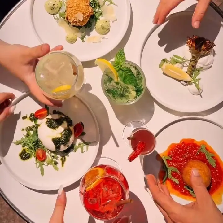 Une table d’antipasti vue du dessus : burrata, arancino, artichaut frit et œuf croustillant, et des mains qui se servent, cocktails en main