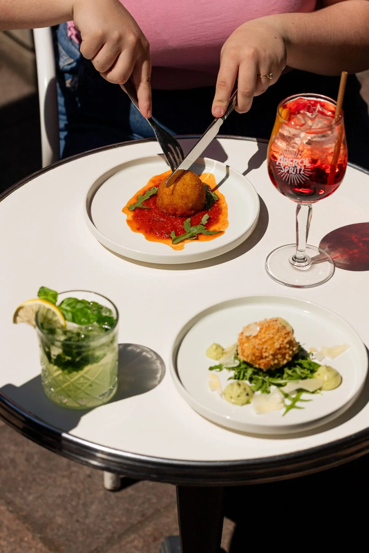 Deux mains coupent un arancini en terrasse, un spritz et un mojito posés sur la table