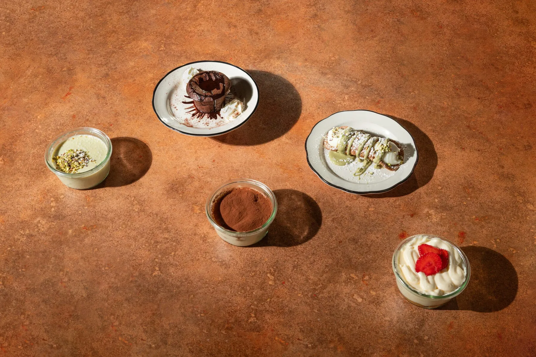Assortiment de desserts vu du dessus : panna cotta à la pistache, moelleux au chocolat, tiramisu, cannolo à la pistache et tiramisu à la fraise