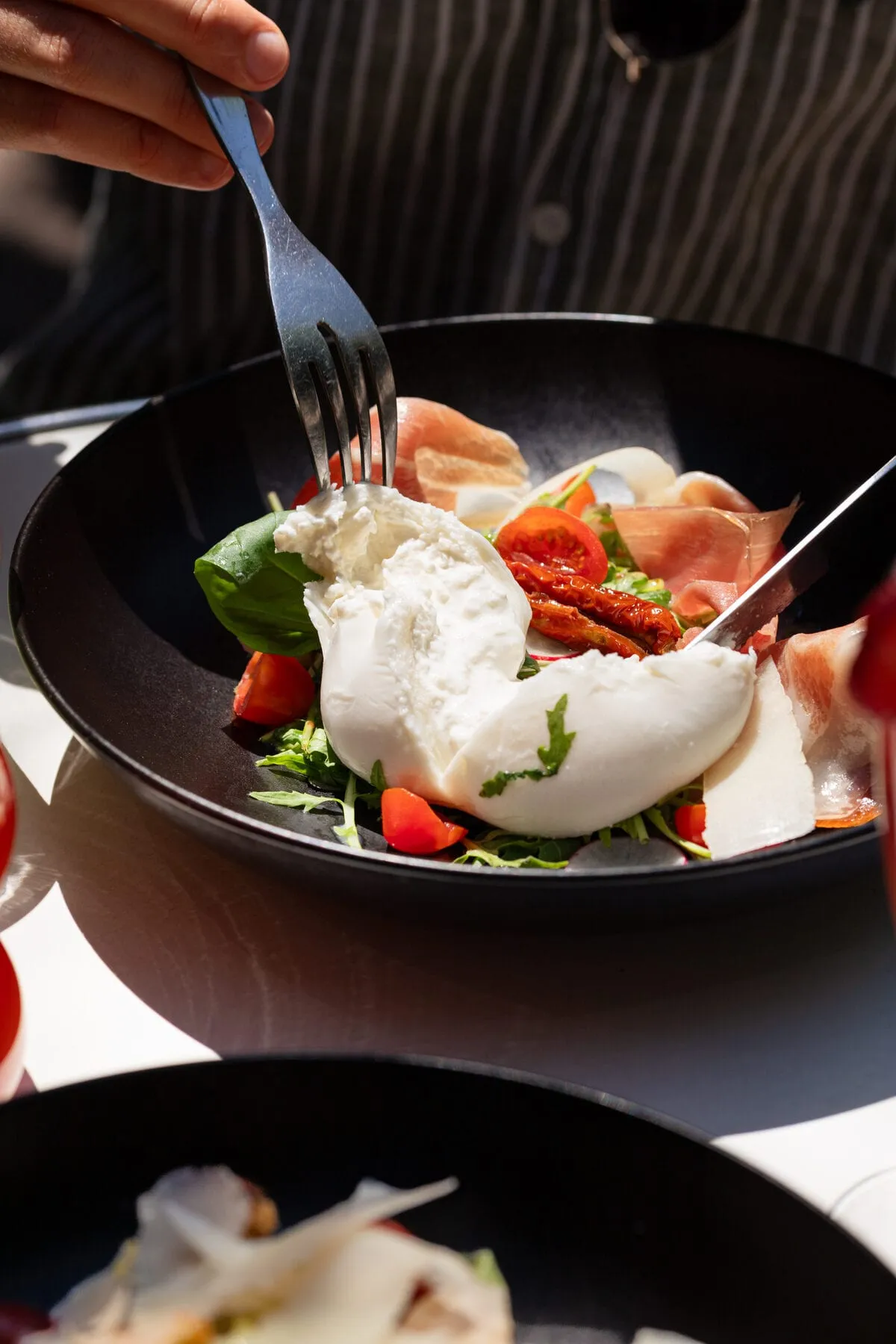 Burrata entamée à la fourchette, jambon de Parme, tomates confites et roquette dans un bol noir