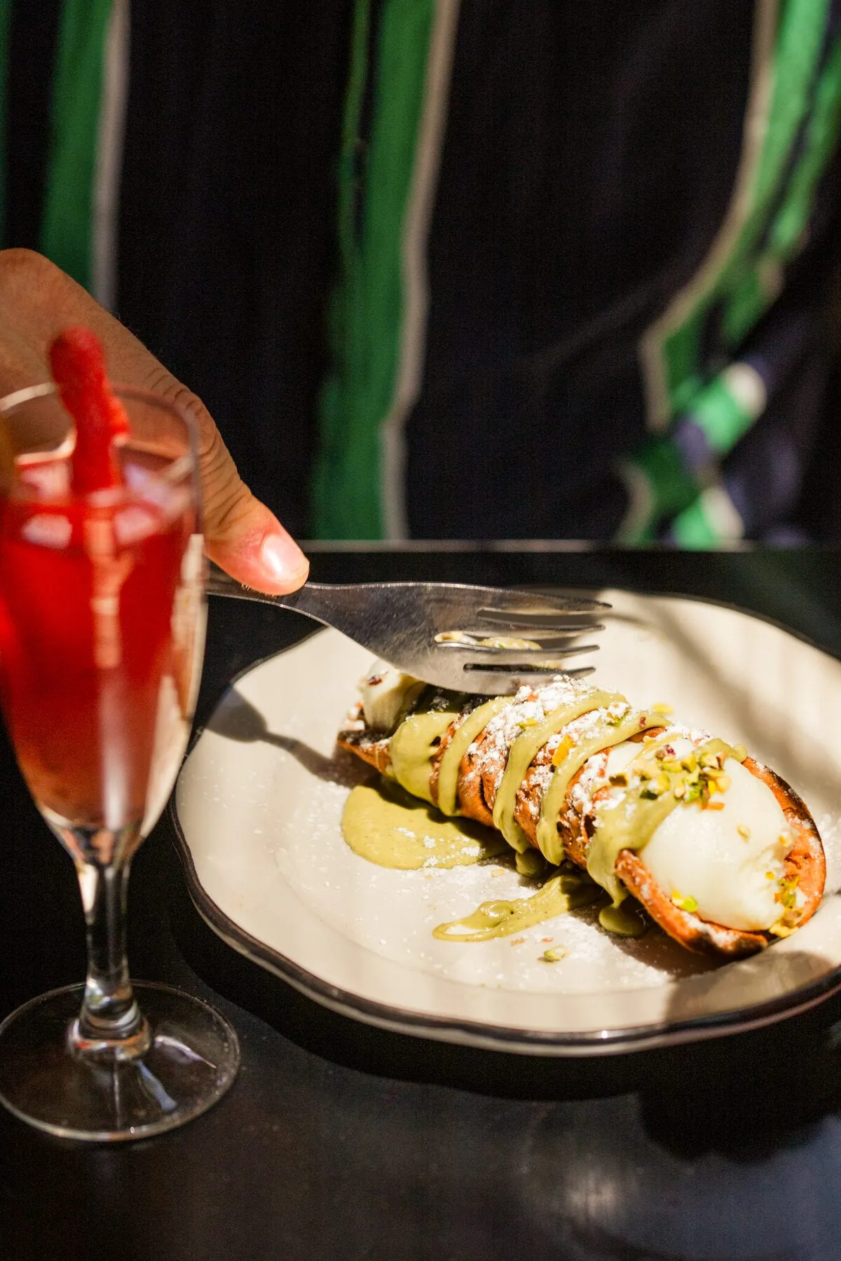 Cannolo nappé de crème de pistache, éclats de pistache et sucre glace, entamé à la fourchette, cocktail à la fraise à côté