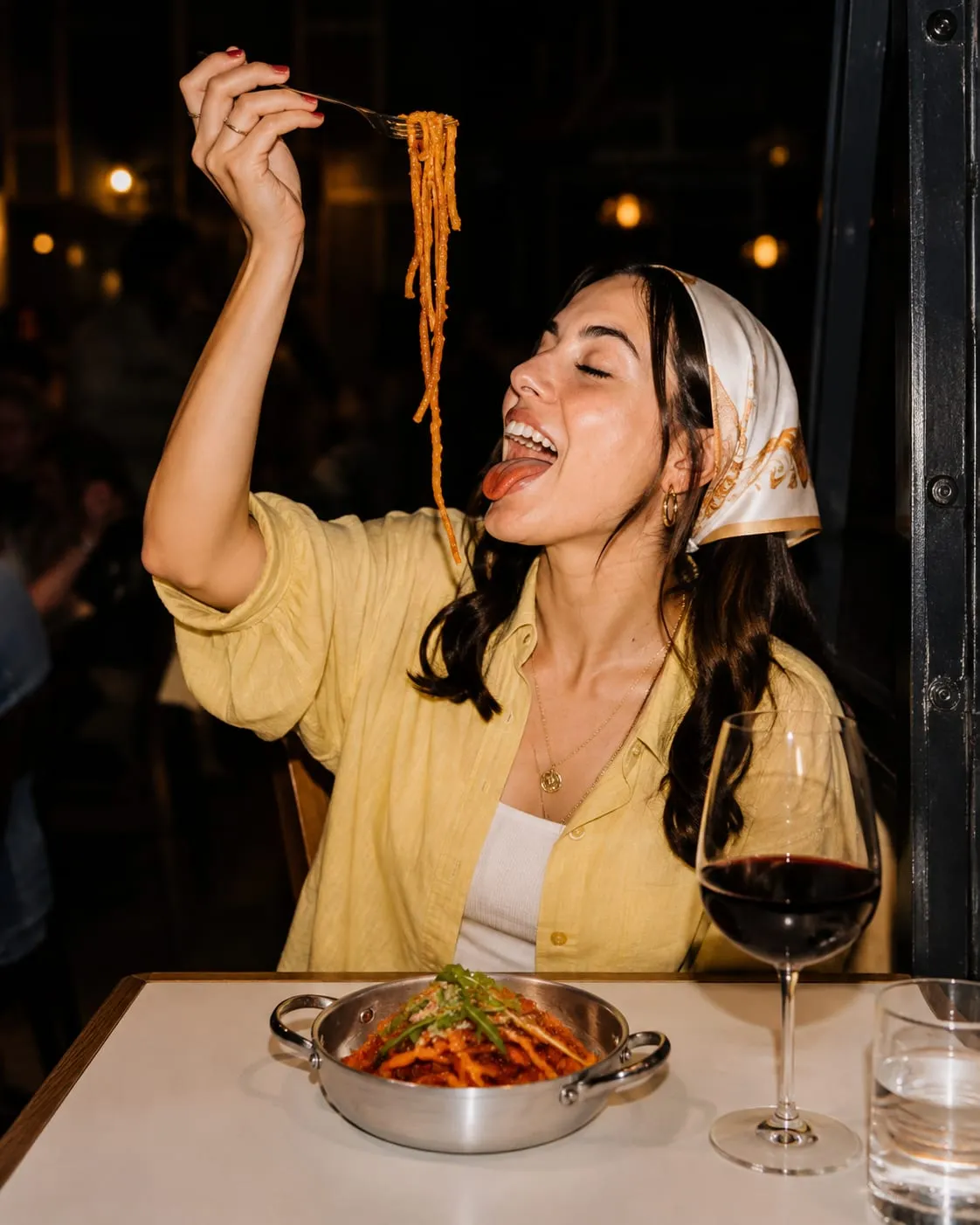 Une cliente en chemise jaune et foulard lève une fourchette de pâtes à la sauce tomate, le soir, en salle, un verre de vin rouge devant elle