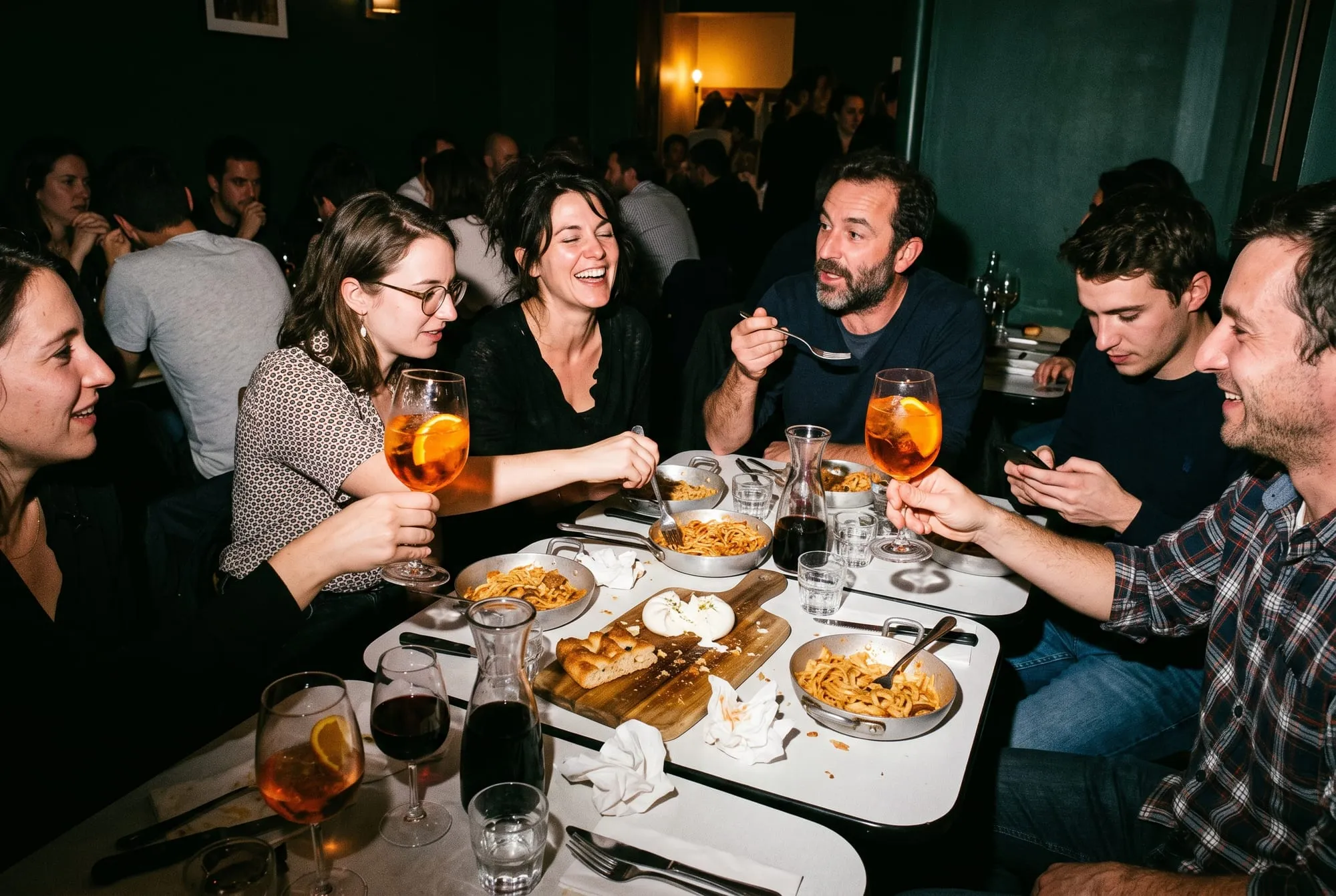 Une bande d’amis rit et bavarde autour de tables rapprochées, le soir, deux spritz levés : pâtes servies au poêlon, burrata ouverte sur une planche de focaccia, carafes de vin rouge